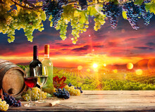 Wine Glasses And Bottle With Grapes And Barrel In Vineyard At Sunset