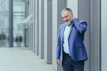 Injured and overtired senior gray-haired businessman, man outside office has severe neck pain, director holding hand massaging neck