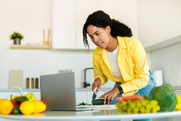 African american woman cooking fresh organic salad at home in modern kitchen, looking at laptop, trying new recipe