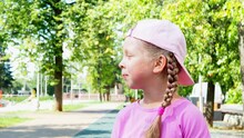 Funny, Cheerful, Little Girl With Braids And In A Pink Cap Looks Attentively To The Side. Portrait Of A Child In Summer In A Sunny Park. A Preschooler Girl Is Watching What Is Happening.