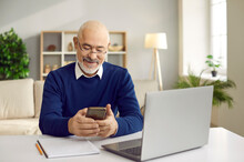 Senior Man Using Mobile Phone. Happy Retired Man In Glasses Sitting At Desk With Laptop And Cellphone, Paying Bills Online, Reading News, Sending Emails, Making Banking Transaction, Calling His Family