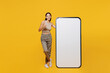 Full body smiling happy young latin woman she wear tank shirt stand near big huge blank screen mobile cell phone with workspace copy space mockup area show thumb up isolated on plain yellow backround.