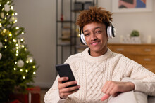 Portrait Of Smiling Man With Wireless Headphones And Phone In Hand, Guy Listens To Music, Podcast, Radio, Audiobook While Relaxing On Couch By Christmas Tree Lights