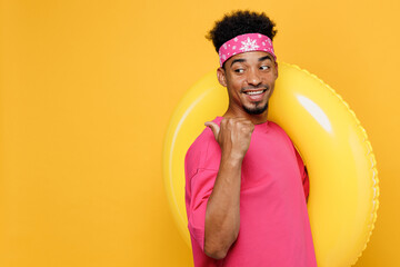Wall Mural - Side view young smiling man he wear pink t-shirt near hotel pool hold inflatable ring point thumb finger aside on workspace area isolated on plain yellow background. Summer vacation sea rest concept.
