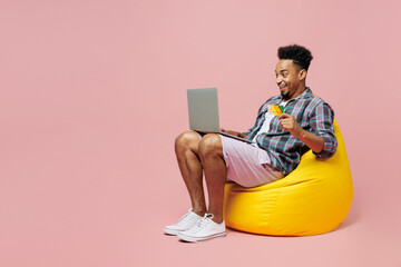 Wall Mural - Full body young man of African American ethnicity wear blue shirt sit in bag chair use mobile cell phone credit bank card shopping online order delivery isolated on plain pastel light pink background.