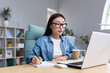 © Liubomir - Distance Learning. Young beautiful Asian female student taking an online exam. Sitting in glasses and jeans at the table at the computer, making notes in a notebook.