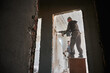 © anatoliy_gleb - Bottom view of two builders deconstructing interior partition. One worker standing on desk with hammer demolishing upper part of wall, another worker by sledgehammer breaking remaining lower part.