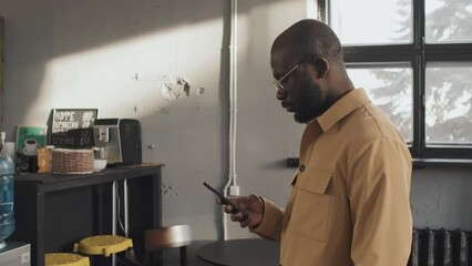 Medium side view of young intelligent Black man using smartphone standing indoors at daytime