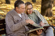 © gpointstudio - Senior couple sitting at the bench in park and looking at old photos