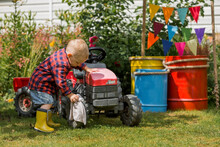 A Cheerful Boy Dressed In A Rustic Style Washes A Tractor With A Rag In The Garden In The Village. Playing In Nature. The Concept Of Raising A Child And Teaching Them To Work.
