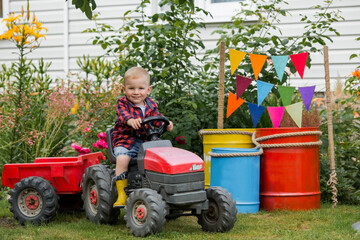 A cheerful boy dressed in a rustic style is sitting on a tractor in a garden in the village. Playing in nature. The child is interested in machinery and machines. The concept of a fun childhood.