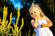 © vita - A pretty little blonde shy girl in a white dress stands in nature among the blooming flowers in a summer park, garden, posing and smiling. Portrait of a 3-5 year old happy female child, kid having fun