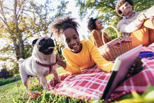Beautiful Young Family Lying On A Picnic Blanket With Their Dog, Enjoying An Autumn Day In Park While Using Digital Tablet.