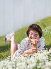 Wall Mural - Woman in colorful sunglasses sniffs clover flowers on lawn in urban park. Nature in town. Relax outdoors after work. Summer vibes.