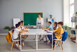 © Studio Romantic - Children having class in cozy classroom interior with green chalkboard. Happy students sitting around big table, reading books, writing in notebooks and talking with teacher. Back to school concept