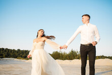 Cheerful Carefree Newlyweds Are Running On The Sand Against The Background Of The Forest, Holding Hands And Looking At Each Other