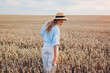 Young woman walking in summer field wearing straw hat and linen shirt holding wheat bundle. Girl touching wheat