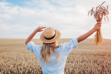 Wall Mural - Back view of happy woman feeling free walking in summer field holding wheat bundle raising arms. Freedom and harmony