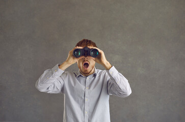 WOW. Studio portrait of funny surprised man holding binoculars and looking open-mouthed ahead standing against grey background. Young guy searches and finds amazing discounts and future seasonal sales