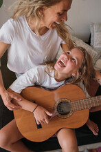 Vertical Photo Of A Blonde Woman Teaching A Girl To Play The Guitar While Looking At Each Other Laughing Sitting On A Couch In A House