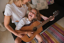 Blonde Woman Teaching A Girl To Play The Guitar While Looking At Each Other Laughing Sitting On A Couch In A House