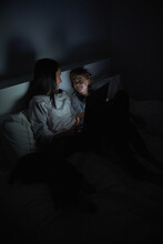 Cheerful Woman And Boy In Nightwear Lying On Bed Under Blanket With Laptop And Watching Movie While Looking At Each Other And Smile During Night Time