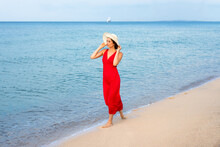 Young Woman In Red Dress And White Hat Walking Along The Sand Beach On A Summer Vacation 