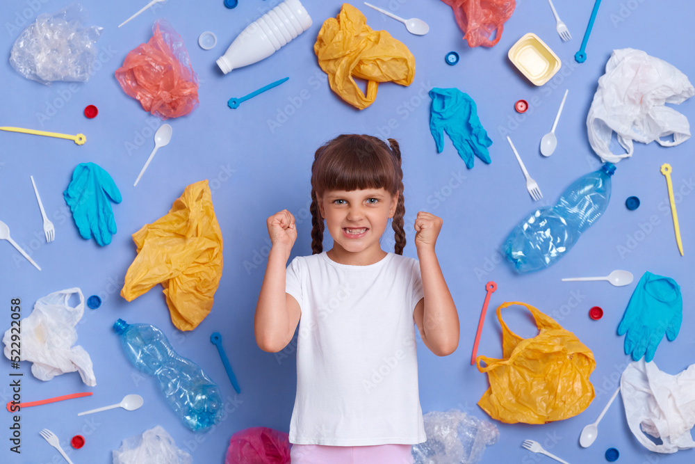 Angry aggressive little girl wearing white t shirt posing against blue wall and much plastic garbage around, clenched fists and screaming with hate, wants to save planet from plastic pollution.