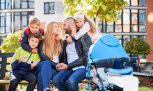 Happy Family - Father, Mother And Children Having Fun Together On Playground. Parents And Kids Sitting On Bench, Hugging Each Other, Looking To Camera. Modern Residential Buildings On Background.