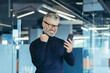 © Liubomir - Successful and satisfied senior businessman celebrating victory gray haired man in glasses working modern office happy reading good news on phone, investor in glasses holding hand up triumph gesture