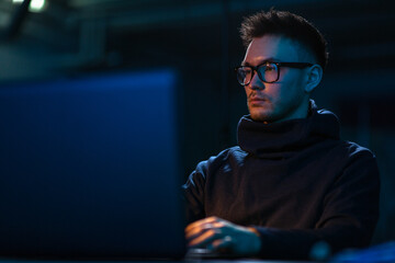 cybercrime, hacking and technology concept - young man in glasses writing code on laptop in dark room