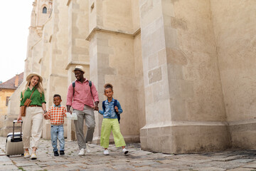 Multiracial family travel together with suitcases, walking in old city centre.