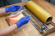 Worker in disposable gloves wraps piece of salmon fillet lying on plastic tray in transparent film.