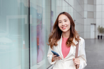 Beautiful asian female businesswoman use smartphone, Walk enjoy smiling while doing commuting in the modern city near office building outside.