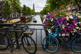 Fototapeta Zachód słońca - Dos bicicletas aparcadas en un puente de Ámsterdam adornado con flores moradas y rosas. En el canal varios barcos y barcas de distintos tamaños. Al fondo, la torre de la iglesia Westerkerk.