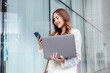 © Kiattisak - Beautiful asian female businesswoman use smartphone and holding laptop computer, Walk enjoy smiling while doing commuting in the modern city near office building outside