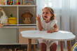 A little girl cries and is naughty sitting at the table in the nursery. Children's tantrums, tears and discontent. Problems of kid's upbringing.