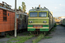 Abandoned Decommissioned Orange And Green Soviet Locomotives Stand On Overgrown Railway In Stock Near Station In Tbilisi, Georgia. Old Railway Machines And Engineering Left Outdoor In City