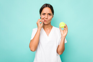 Wall Mural - Young hispanic physiotherapy holding a tennis ball isolated on blue background with fingers on lips keeping a secret.