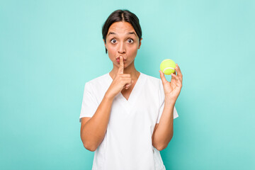 Wall Mural - Young hispanic physiotherapy holding a tennis ball isolated on blue background keeping a secret or asking for silence.