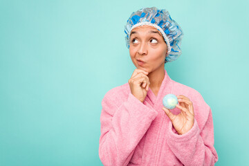 Wall Mural - Young hispanic woman holding bathtub ball isolated on blue background looking sideways with doubtful and skeptical expression.