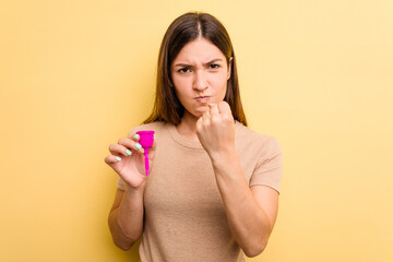 Young caucasian woman holding a menstrual cup isolated on yellow background showing fist to camera, aggressive facial expression.