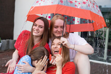 Group Of Young Laughing Stunning Women Sitting On Wooden Veranda, Hiding From Rain Under Umbrella With Dots, Having Fun.