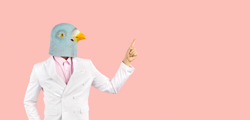 Strange guy with funny bird head points his index finger at blank copy space on right side. Man in suit and wacky pigeon mask showing something on solid pastel pink text copyspace banner background