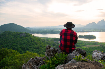 Wall Mural - A young tourist in a plaid shirt and black hat sits alone on the mountaintop overlooking the beautiful view of the lake and  sunset. He felt happy, enjoy and relaxed to watch the sunset from mountain.