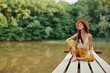 © SHOTPRIME STUDIO - Hippie eco-activist woman traveler sits on a bridge by a lake with her arms outstretched with a hat and smiling sincerely