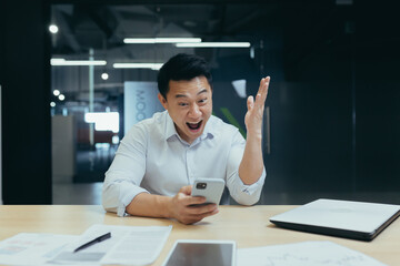 Successful and happy asian businessman man celebrating victory sitting and working in modern office at desk, celebrating victory looking at camera and joyfully shouting holding mobile phone.