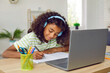© Studio Romantic - Happy elementary school child studying online. African American student girl in headphones sitting at desk with laptop computer at home, writing in notebook, having remote class or listening to music