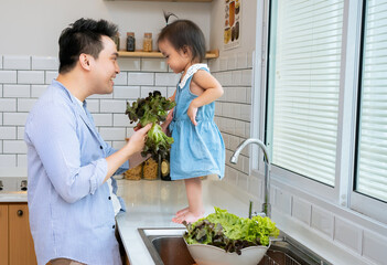 Sweet little Asian girl helping dad to prepare dinner, making salad from fresh vegetables. African American young father teaching kid to cook in home kitchen at table with organic food ingredients.