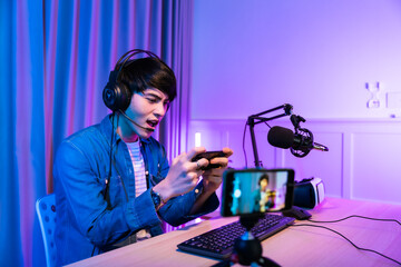 Wall Mural - Playing video games on smartphone. Young asian handsome man sitting on chair holding cellphone in his hand. Exited streamer wearing headset  in neon room .Esport streaming game online.